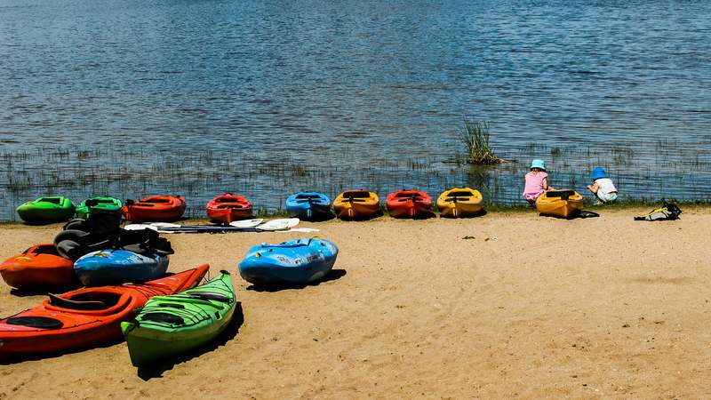 Overhead view of a brightly colored recreational kayak on calm blue water beside a coastal rental dock with insurance document icon overlaid